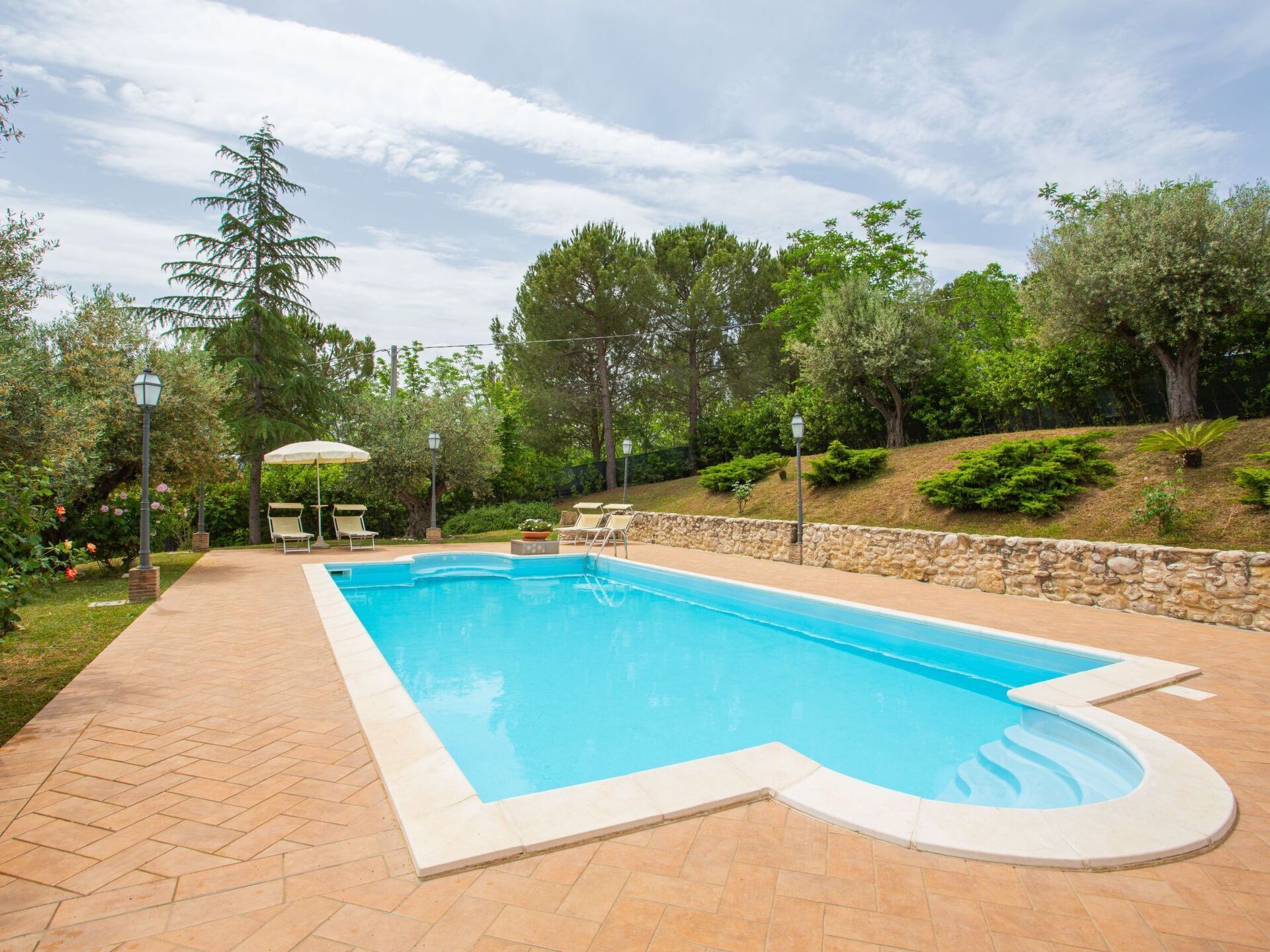 A rectangular swimming pool with a stone wall and tiled deck, surrounded by greenery and trees.