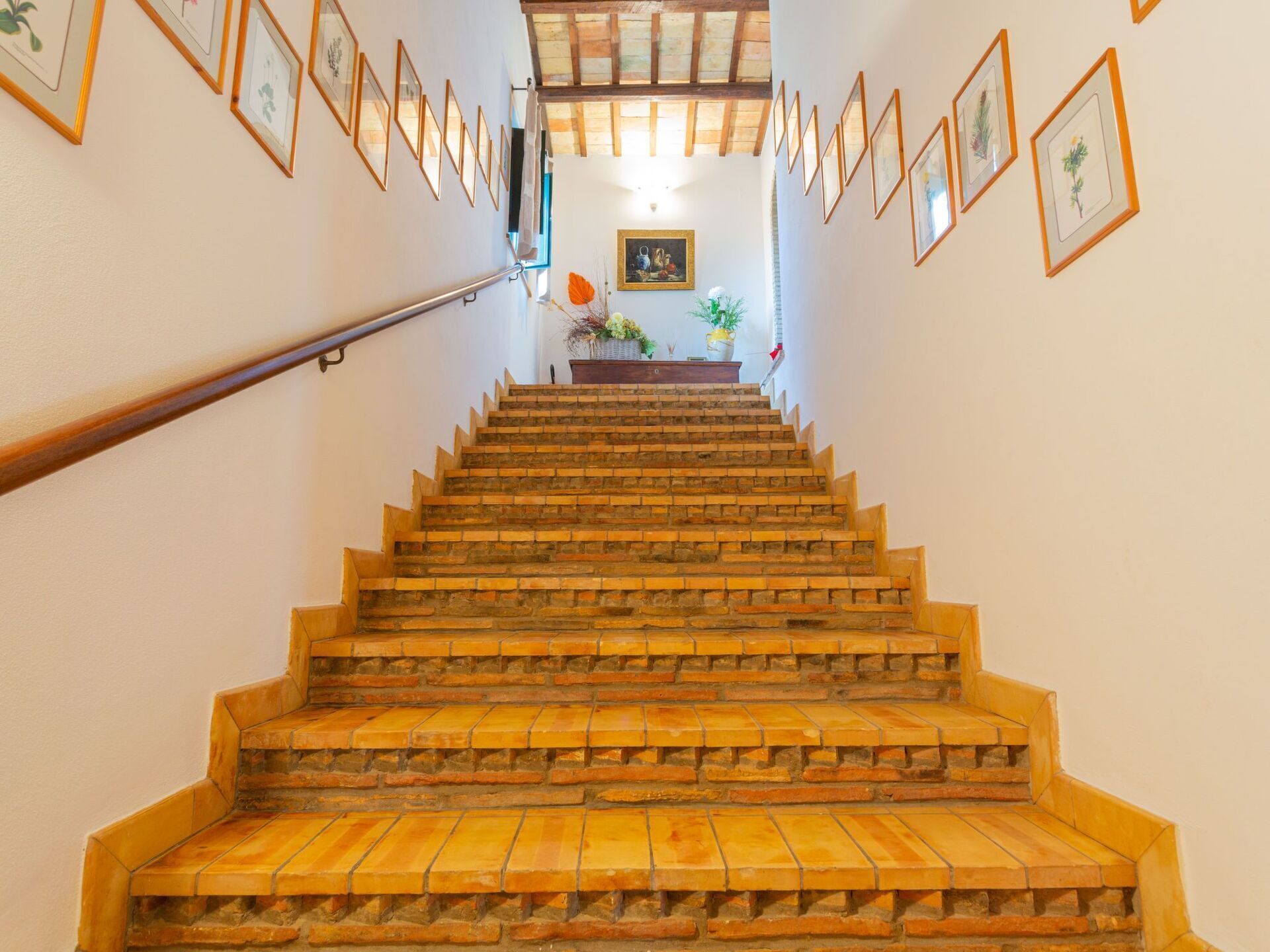 A staircase with a handrail, leading to a landing with a framed picture and a vase of flowers.