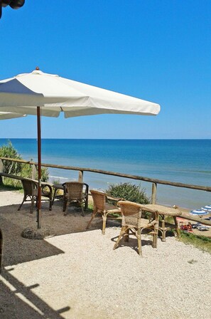 Et siddeområde ved stranden med fletstole og en hvid parasol, med udsigt over havet.