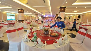 Une salle de banquet avec des tables rondes préparées pour un événement formel, chacune étant recouverte d’une nappe blanche, ornée d’un ruban rouge et agrémentée d’une vaisselle soigneusement disposée.