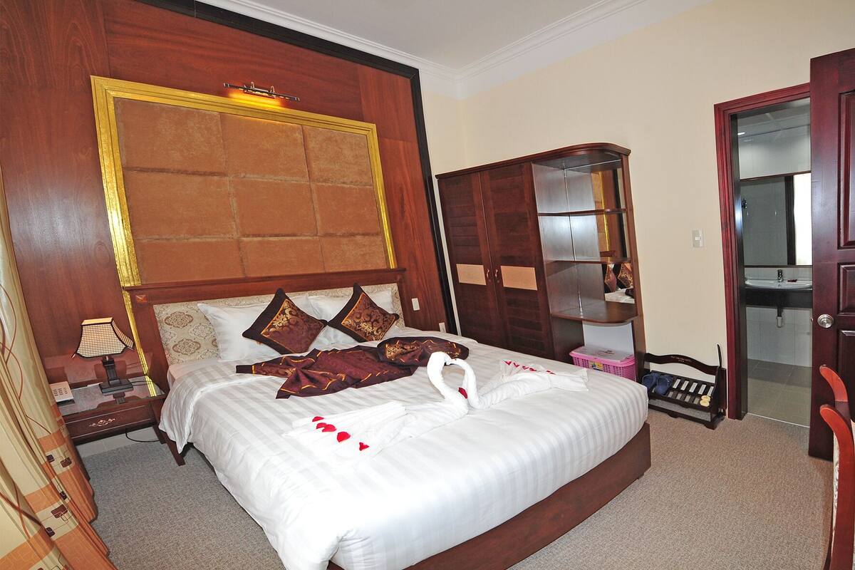 Une chambre d’hôtel avec un grand lit, une tête de lit en bois, une armoire, un miroir et un petit banc.