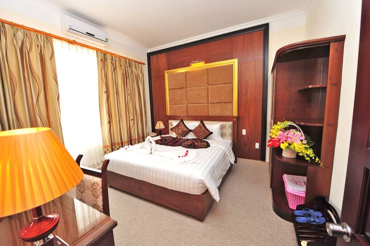 Une chambre d’hôtel avec un grand lit, une tête de lit en bois, une table de chevet avec une lampe, un bureau avec une chaise, une armoire et une étagère ornée de fleurs.