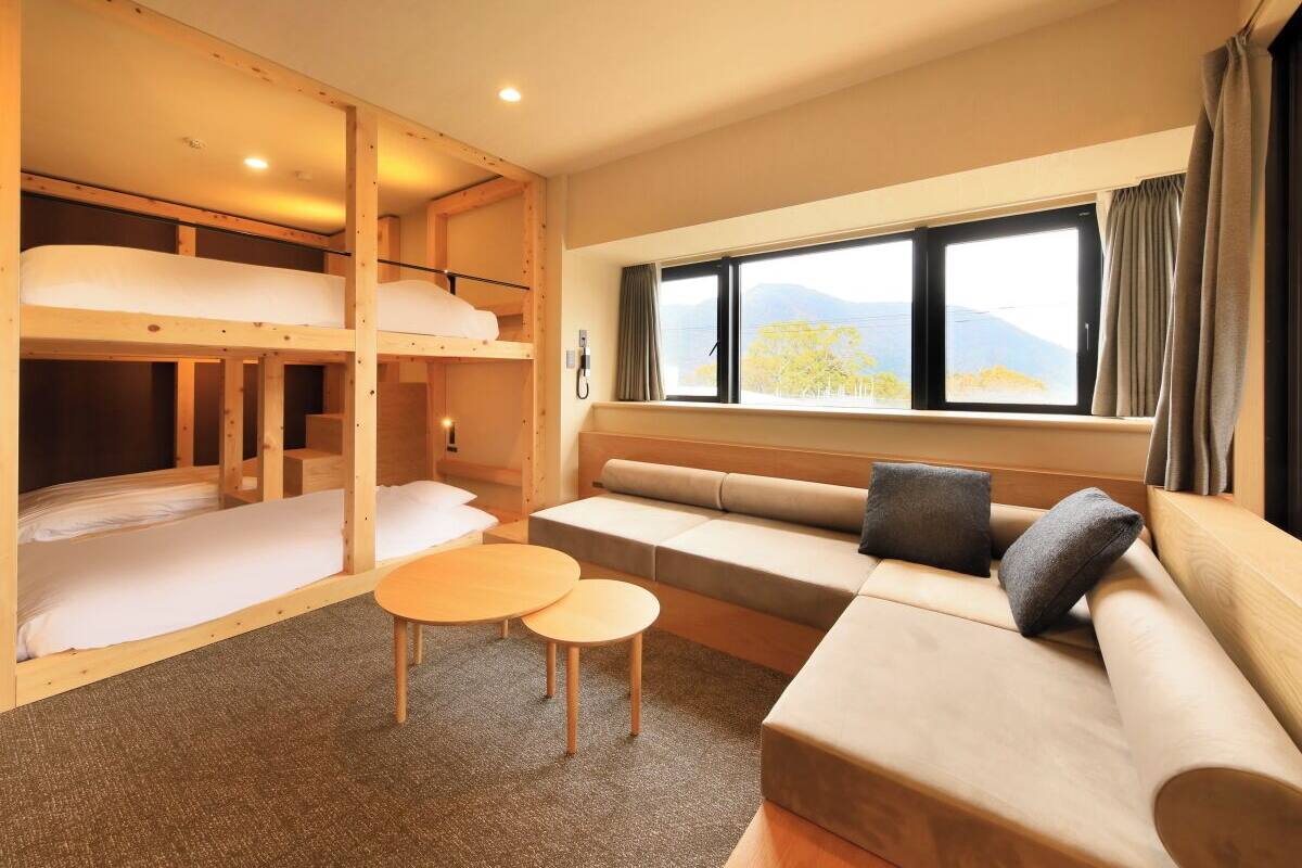 A compact hotel room with bunk beds, a sofa, a small table, and a window with a view of mountains.