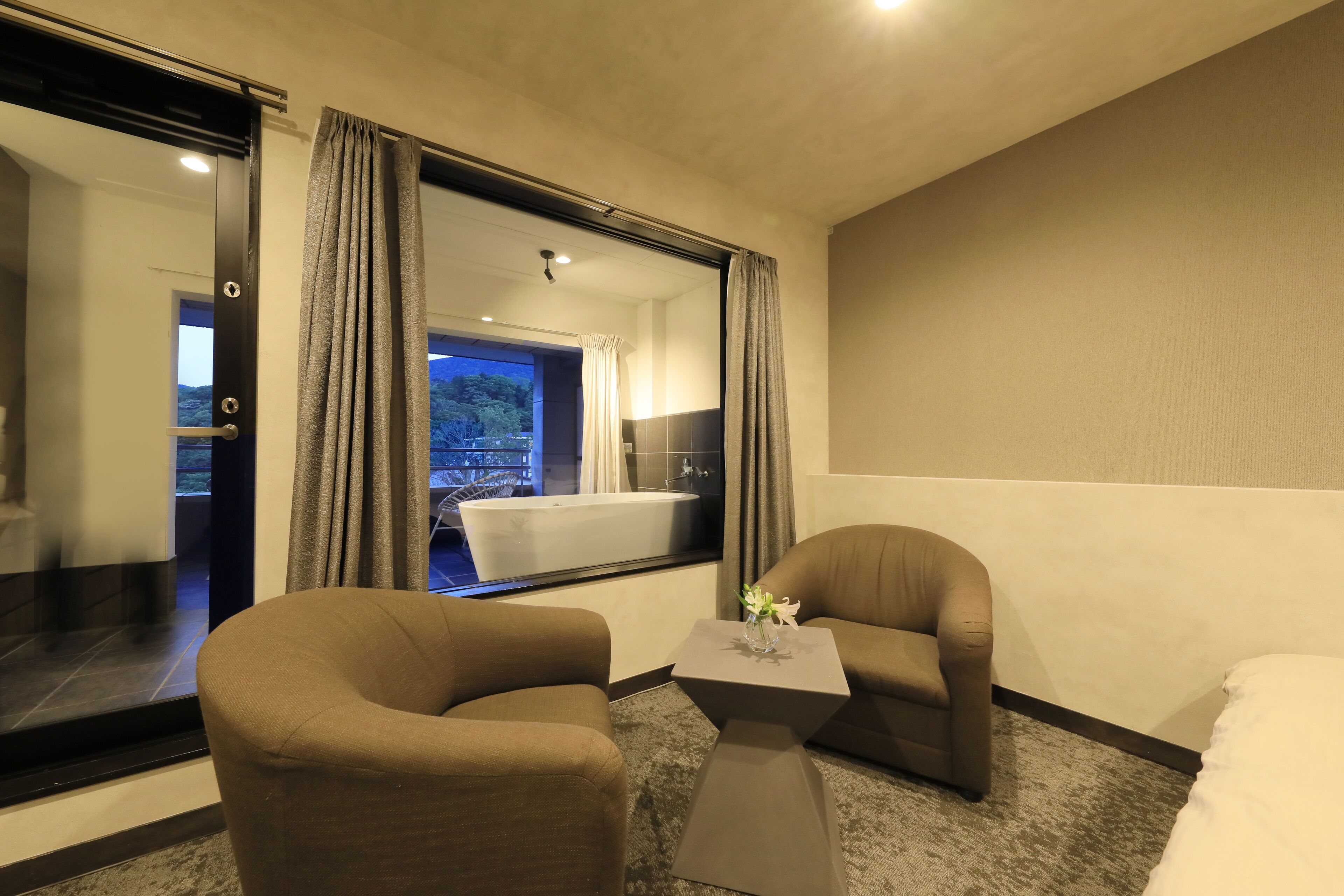 Una habitación de hotel con un ventanal grande, dos sillones, una mesita y una cama.