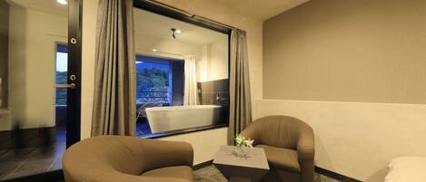 Una habitación de hotel con un ventanal grande, dos sillones, una mesita y una cama.