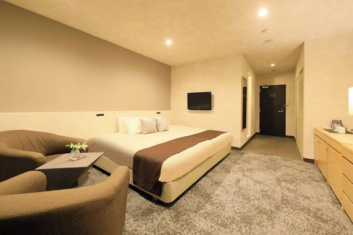 A hotel room with a bed, a desk, a chair, a TV, and a bathroom visible in the background.