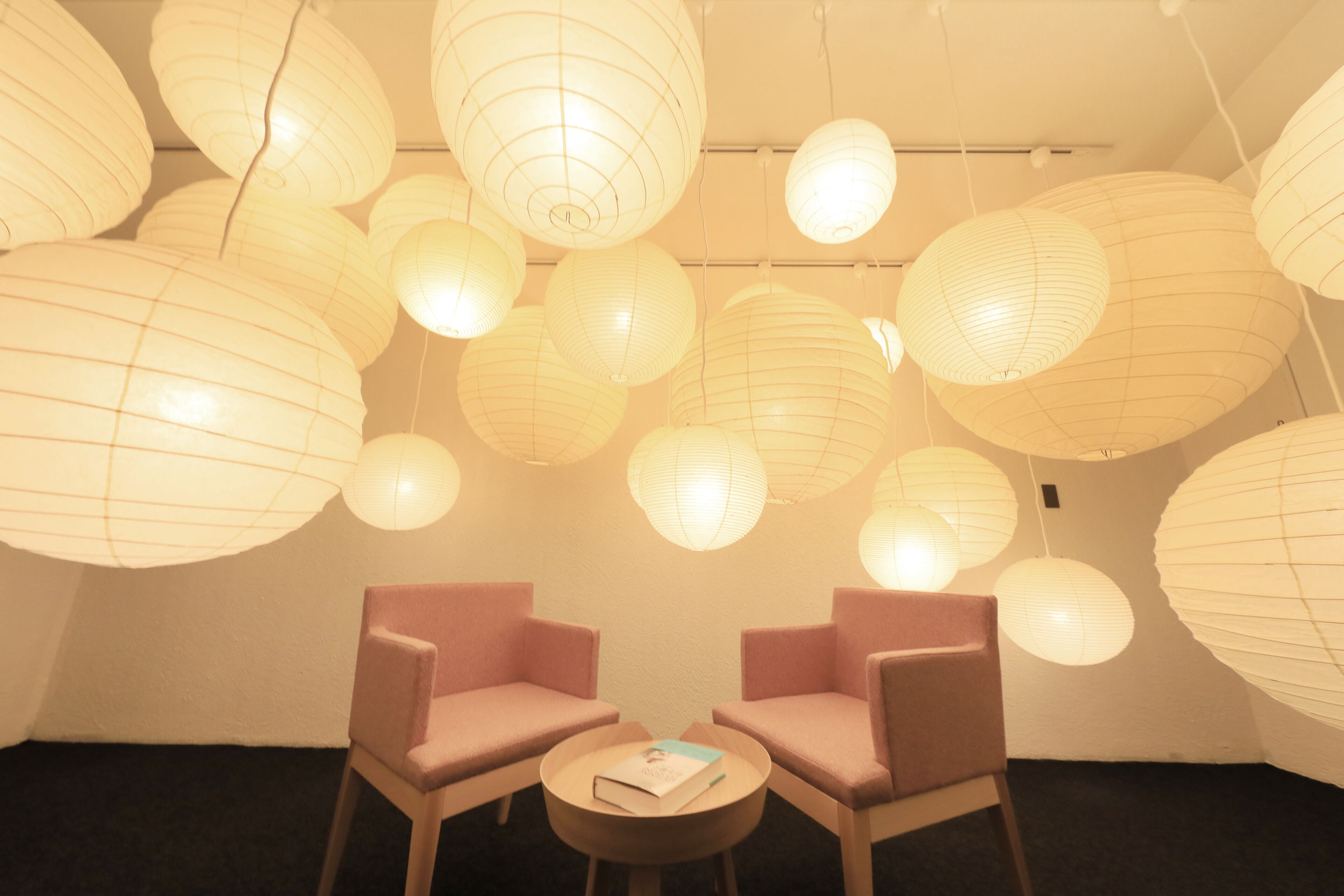 A room with multiple hanging paper lanterns, two pink chairs, and a small round table with a book.