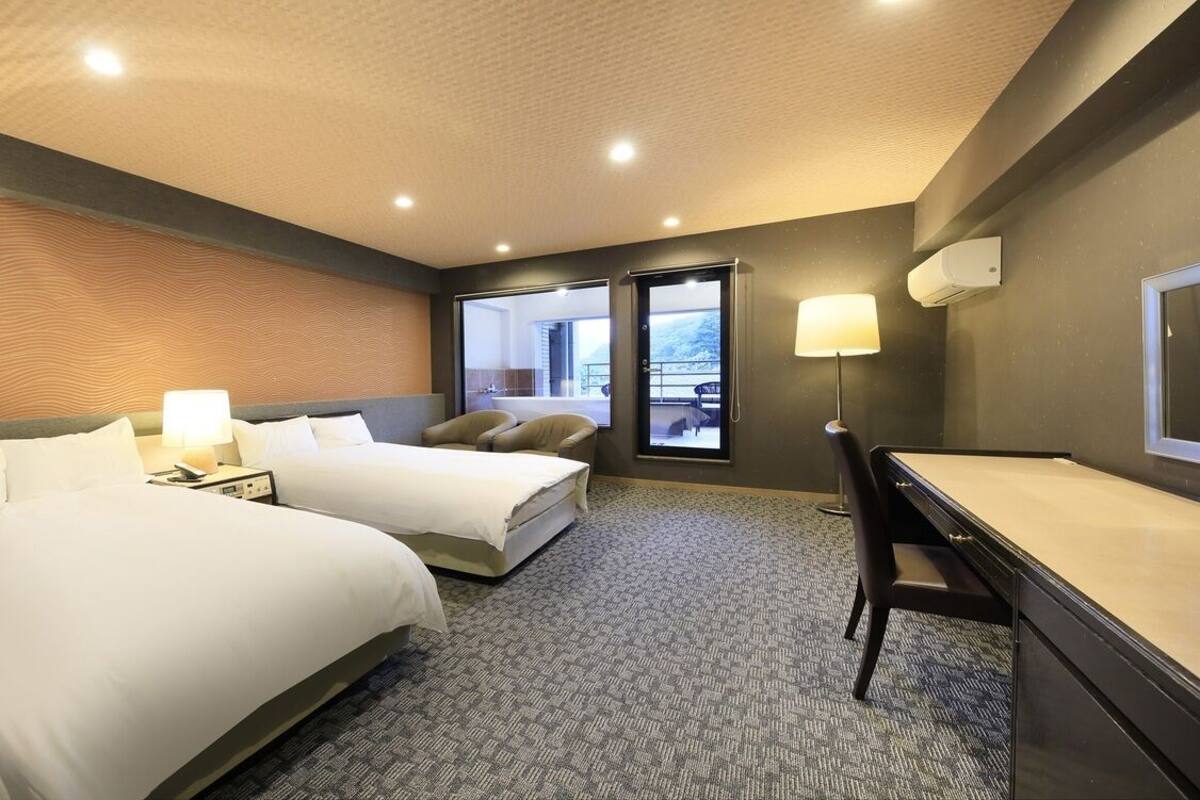 A hotel room with two beds, a desk, a chair, a window with a view, and a ceiling with recessed lighting.