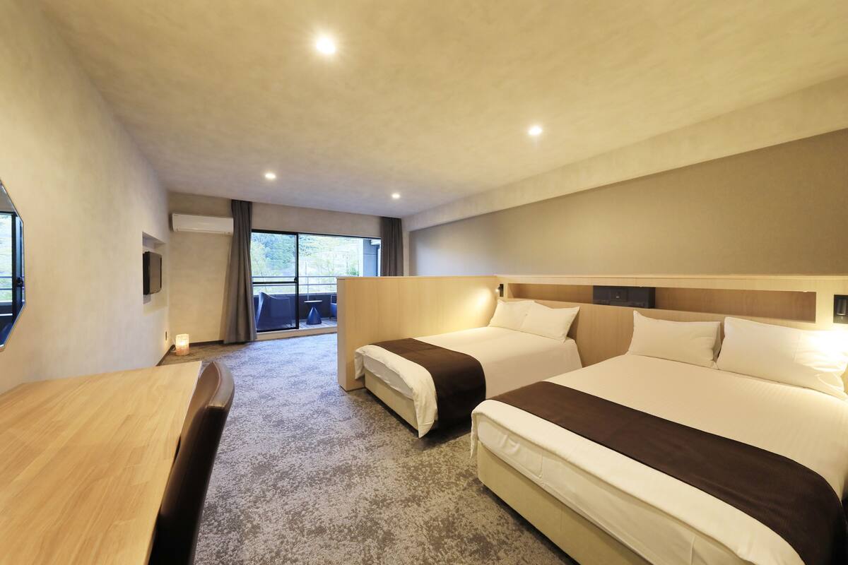 A modern hotel room with two beds, a wooden desk, and a large window providing a view of the outdoors.