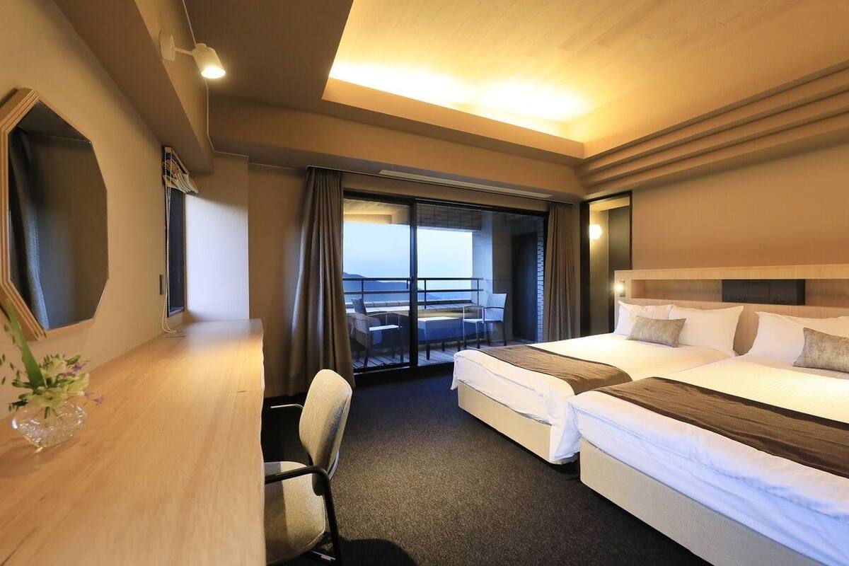 A hotel room with two beds, a desk, a chair, and a large window with a view.