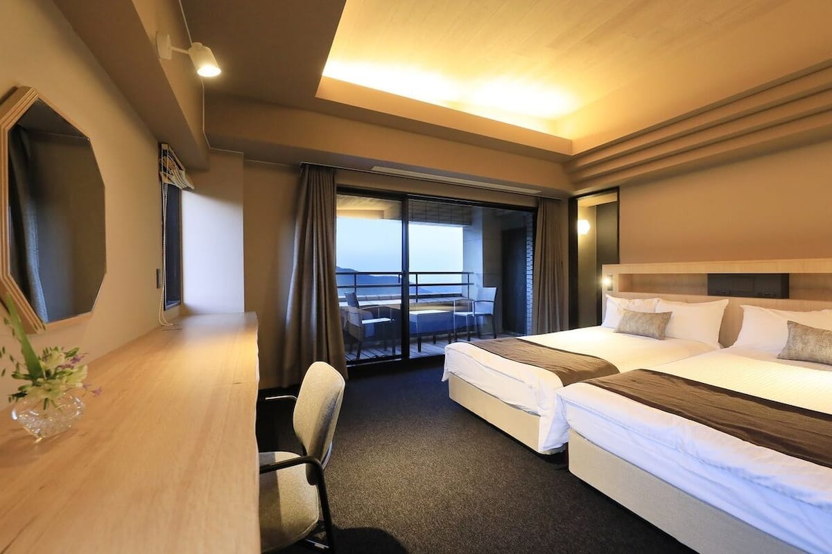 Habitación de hotel con dos camas, un escritorio, una silla y un ventanal con vistas.