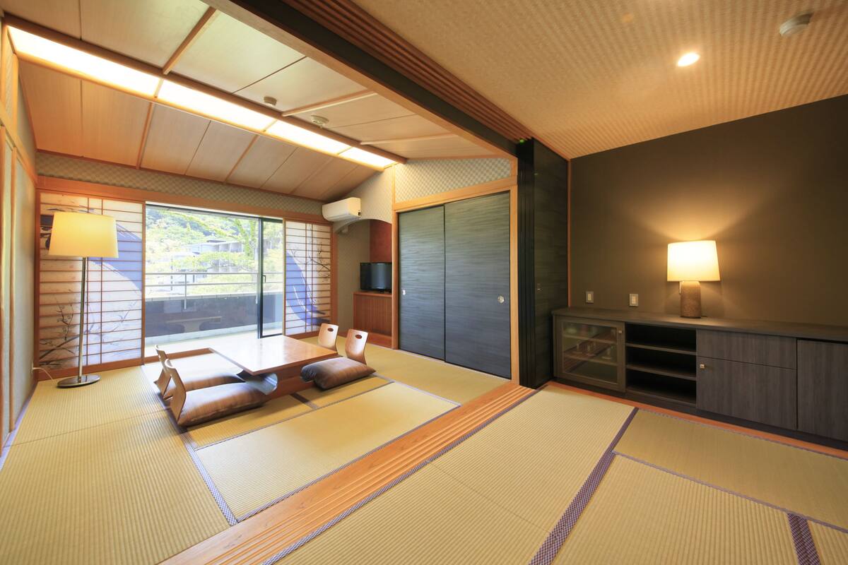A traditional Japanese room with tatami flooring, a low table with cushions, a sliding door, and a TV on a cabinet.