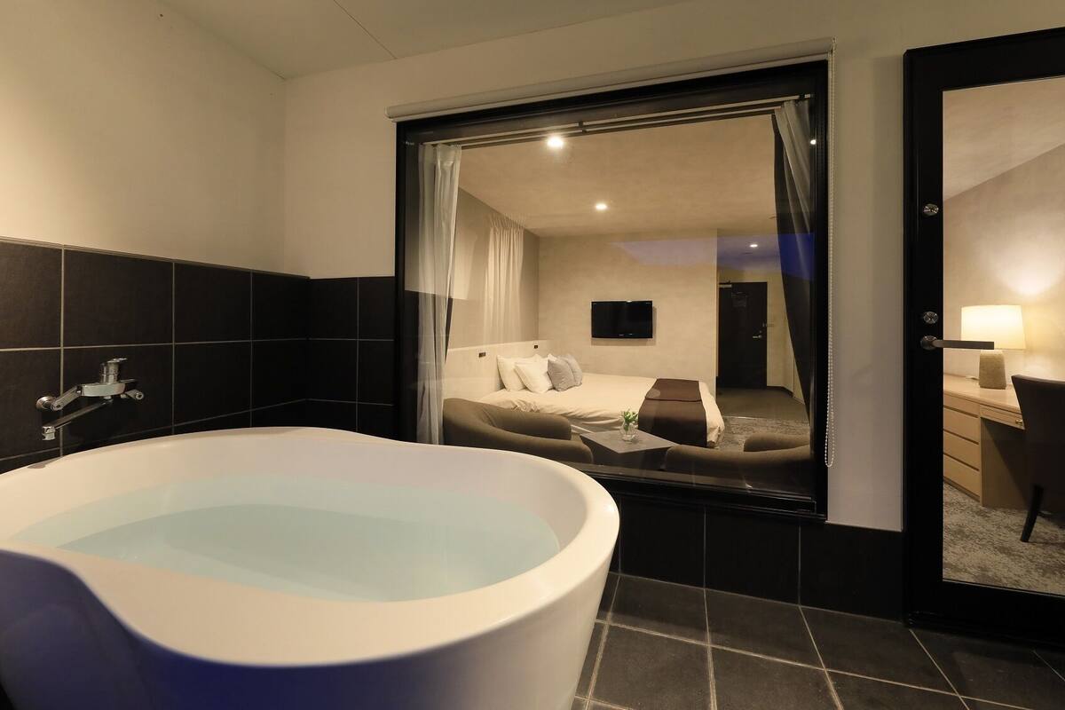 A modern bathroom with a freestanding bathtub, a shower, and a large mirror reflecting a bedroom with a bed and a desk.