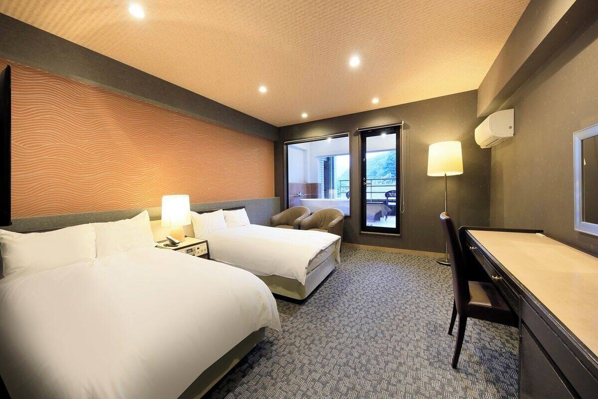 A hotel room with two beds, a desk, a chair, a lamp, and a view of the outdoors.