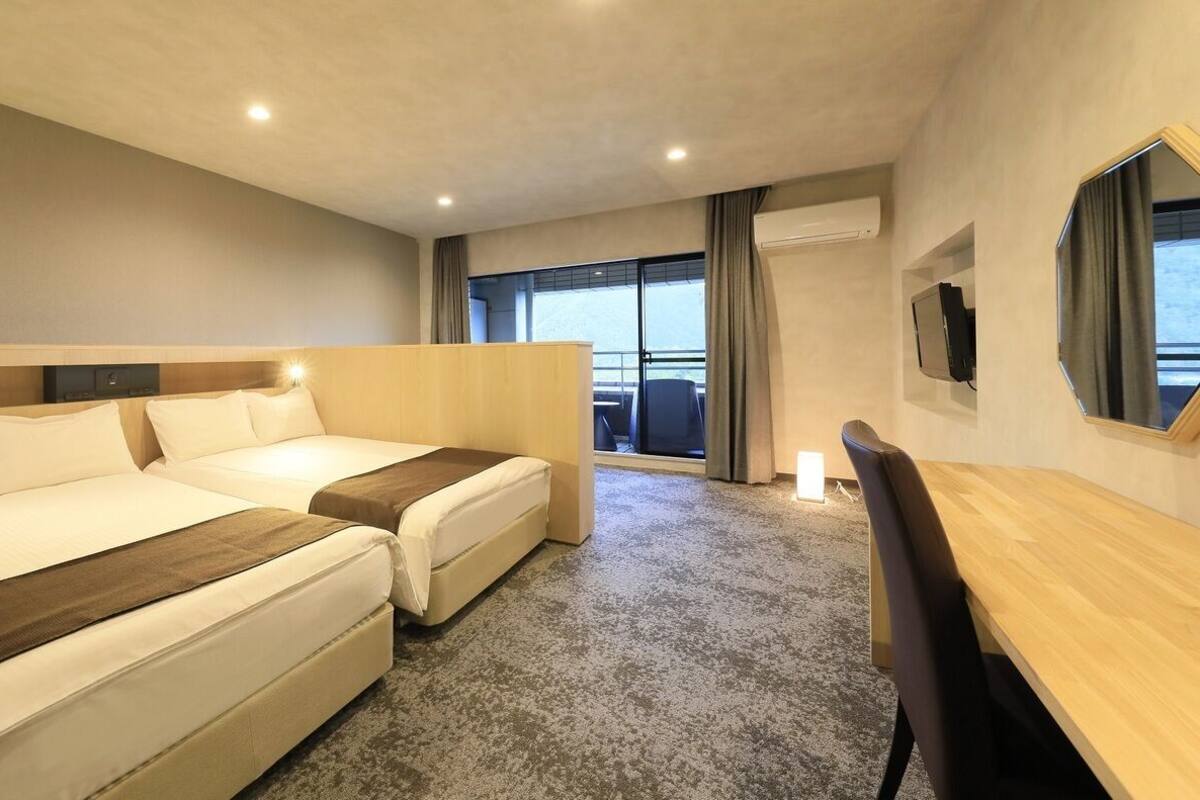 A hotel room with two beds, a desk, a chair, a TV, and a large window with a view.