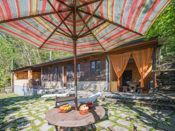 Una cabaña rústica con fachada de piedra, una terraza de madera y una gran sombrilla a rayas que proporciona sombra.
