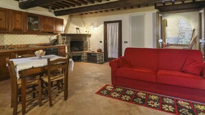 A cozy living room with a red sofa, a dining table with chairs, a fireplace, and a kitchen area with wooden cabinets and a stone fireplace. - Lovely apartment for 6 guests with private pool, WIFI, A/C, TV, terrace and pets allowed (Lugliano)