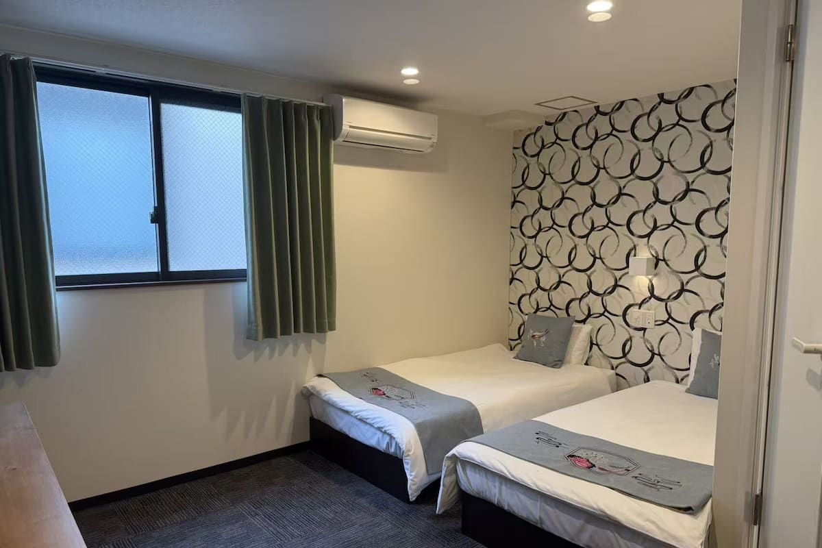 A hotel room with two beds, a window with curtains, an air conditioning unit, and a patterned wall.