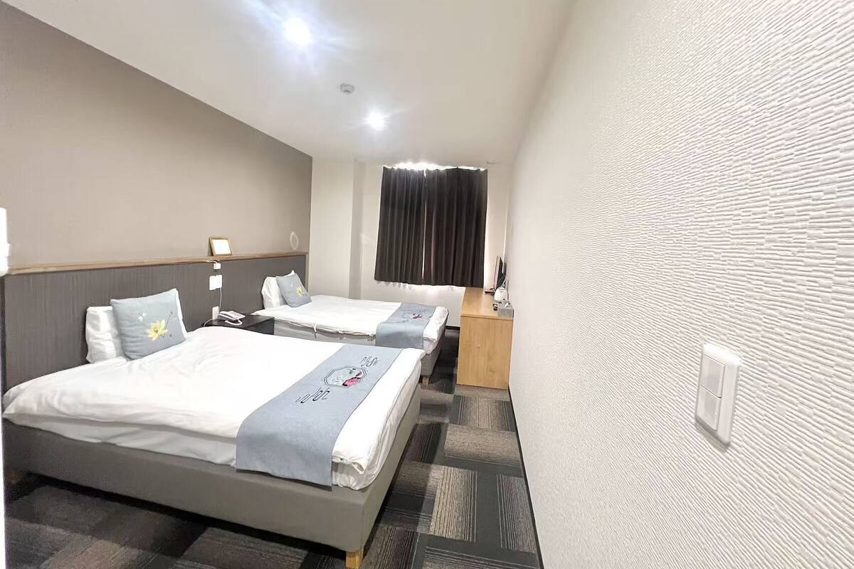 A hotel room with two beds, a desk, a chair, a window with curtains, and a textured wall.