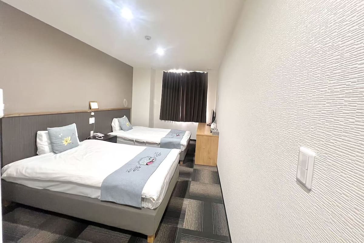 A hotel room with two beds, a desk, a chair, a window with curtains, and a textured wall.