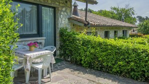 Il s’agit d’un bâtiment en pierre, doté d’un toit en tuiles, d’une petite fenêtre et d’une porte. On y trouve une table avec des chaises et un panier de fleurs à l’extérieur.