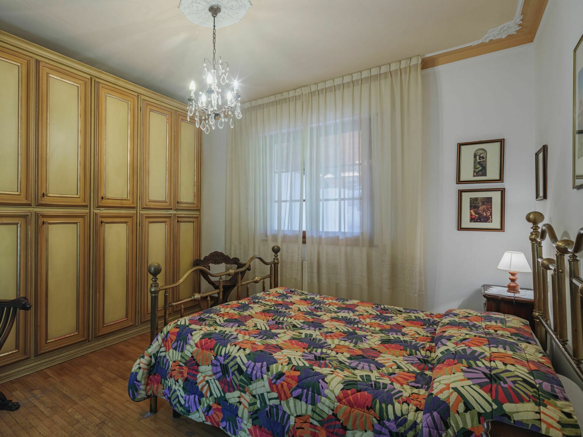 A bedroom with a large bed, wooden wardrobe, chandelier, and a window with sheer curtains.