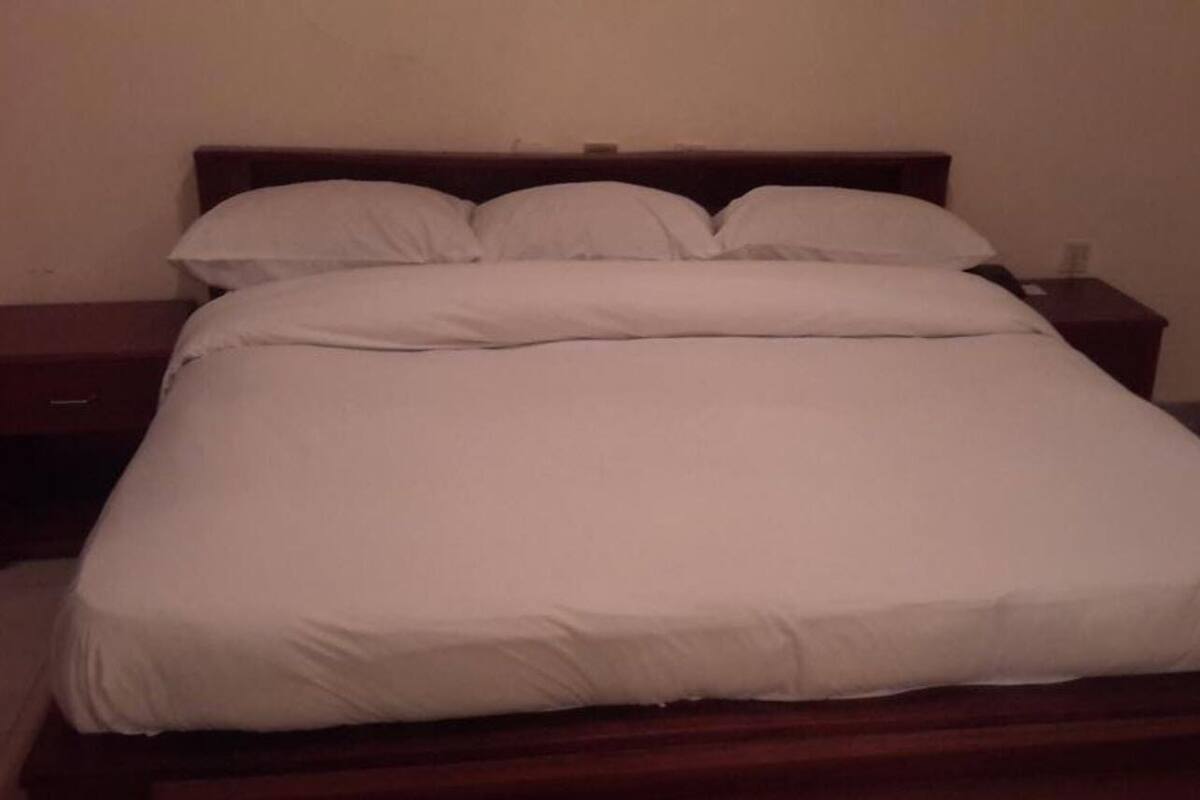 Quarto de hotel padrão com uma cama de solteiro, mesa de cabeceira e telefone fixado na parede.