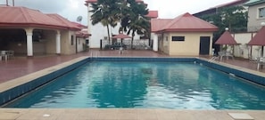 A swimming pool with a tiled deck, surrounded by buildings with red roofs and palm trees. - Owerri Hotel Plaza (Owerri)