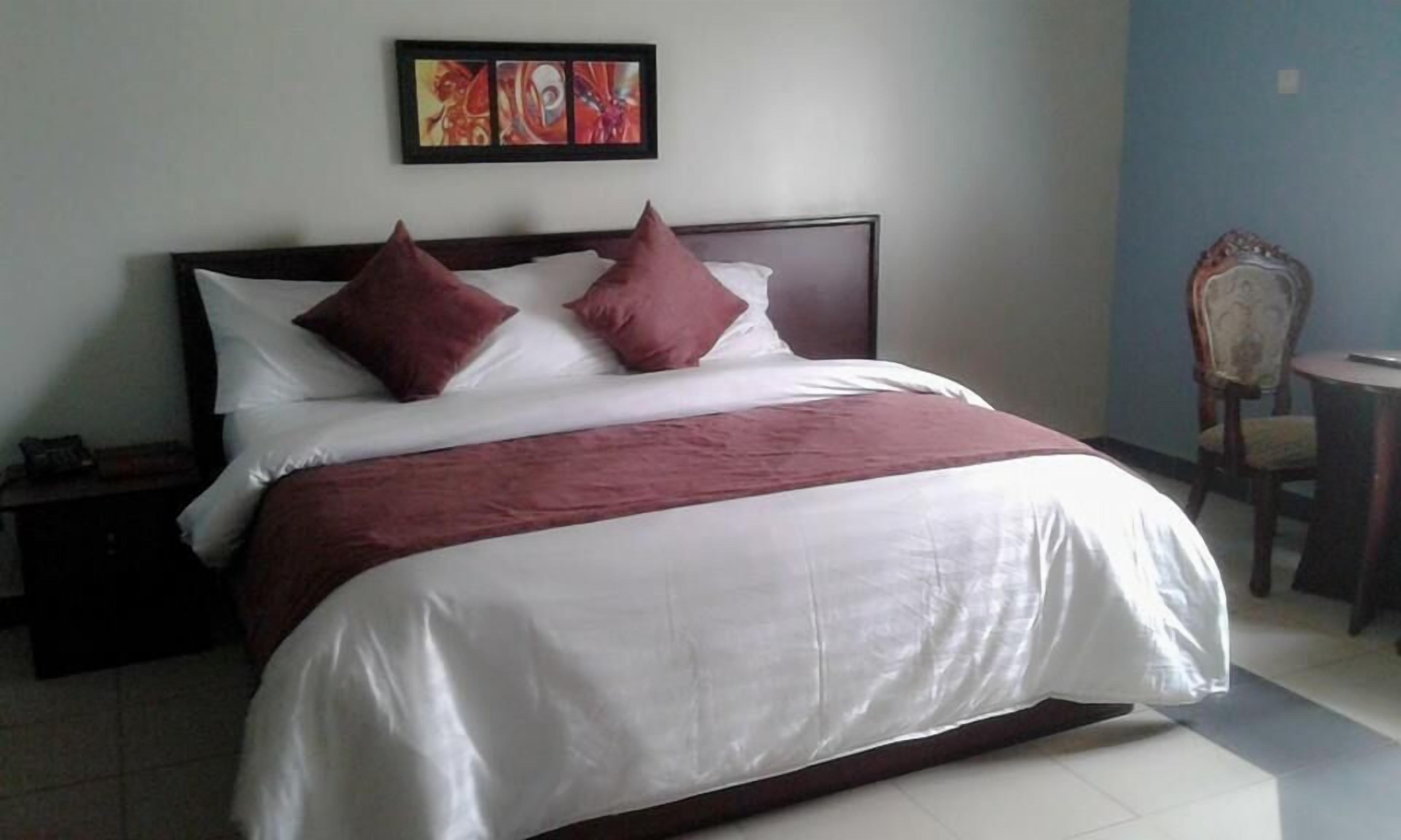 A neatly made bed with a white and maroon bedspread, two maroon pillows, and a wooden headboard. There is a small table with a chair to the right, and a painting above the bed.