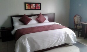 A neatly made bed with a white and maroon bedspread, two maroon pillows, and a wooden headboard. There is a small table with a chair to the right, and a painting above the bed. - Owerri Hotel Plaza (Owerri)