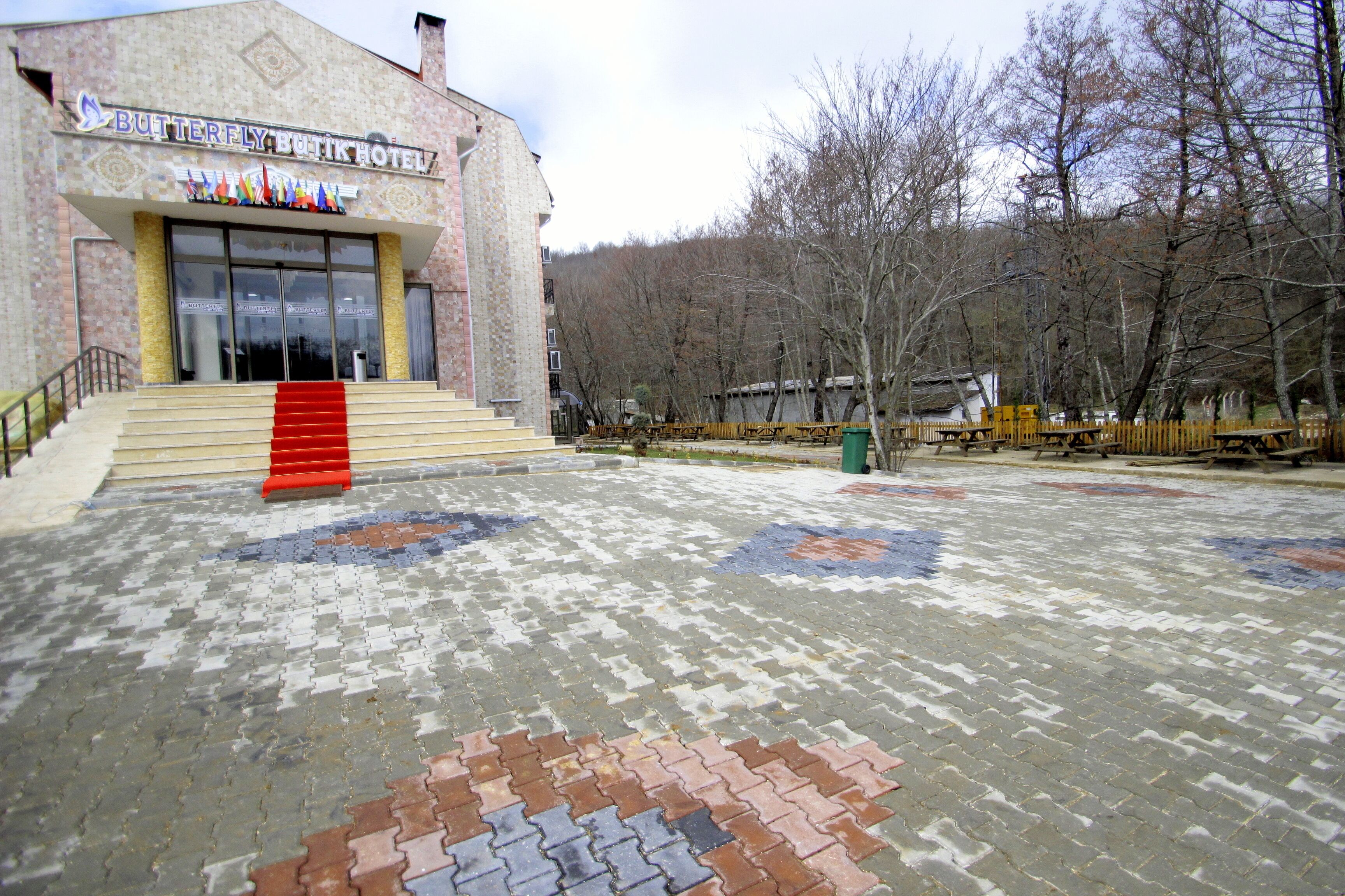 Entrada de hotel con alfombra roja que conduce a las puertas, pavimento estampado y un paisaje boscoso de fondo.