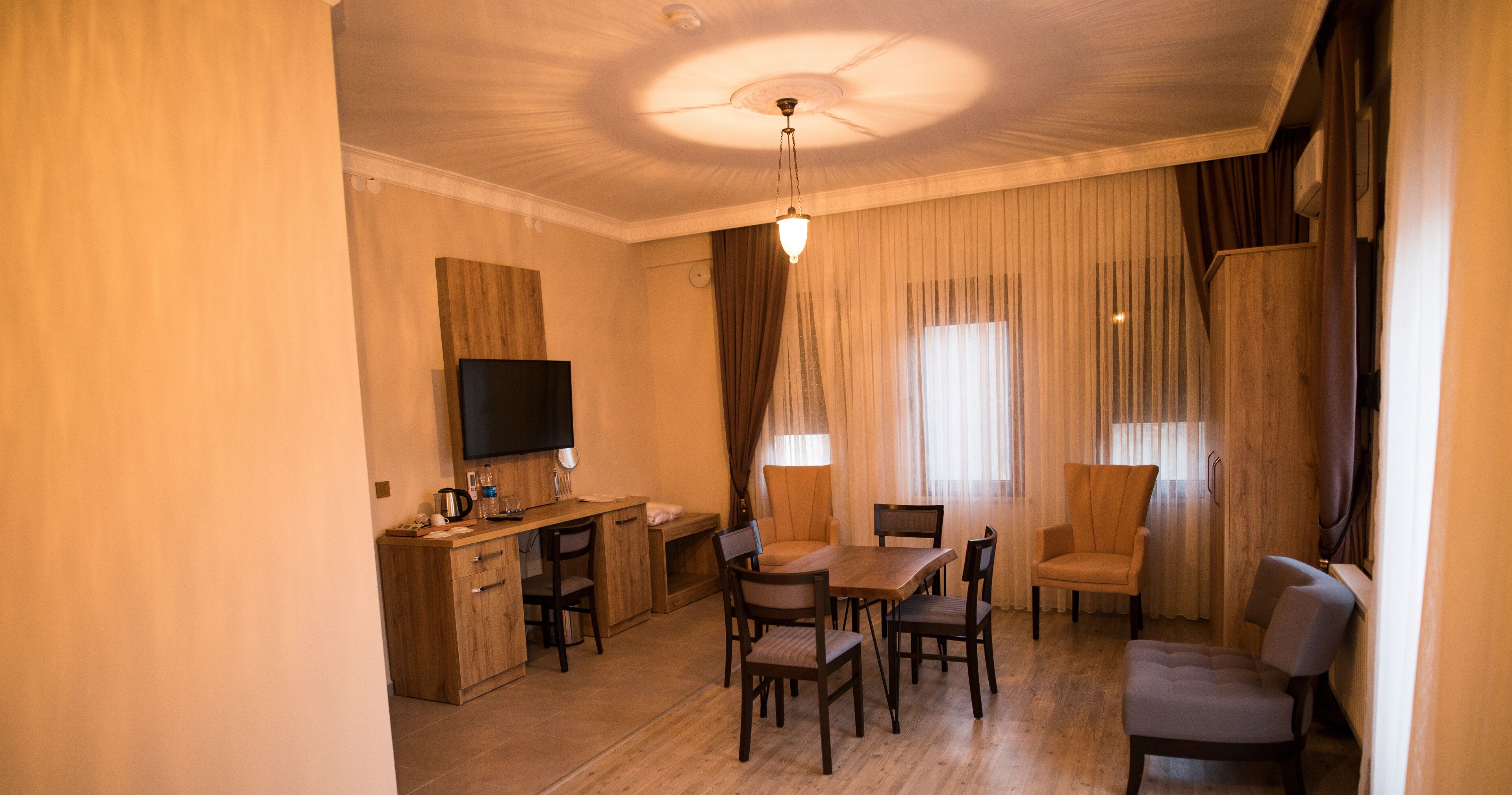 A hotel room with a wooden desk, a flat-screen TV, a dining table with chairs, and a large window with sheer curtains.
