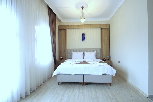 Una cama bien hecha en una habitación de hotel, con cabecera de madera, una mesita de noche con lámpara y un ventanal con cortinas translúcidas.