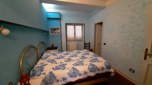 A bedroom with a bed, a chair, a small table, a window with curtains, and a radiator.