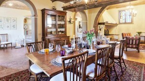 Uma sala de jantar com uma mesa de madeira posta para seis pessoas, rodeada por cadeiras. Há um armário com portas de vidro, um candeeiro de cristal e um tapete estampado no chão.