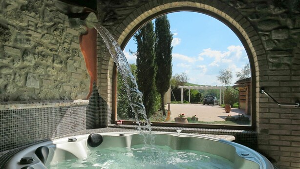 Una vasca idromassaggio con acqua che scorre, affacciata su un muro in pietra, un giardino e un edificio.