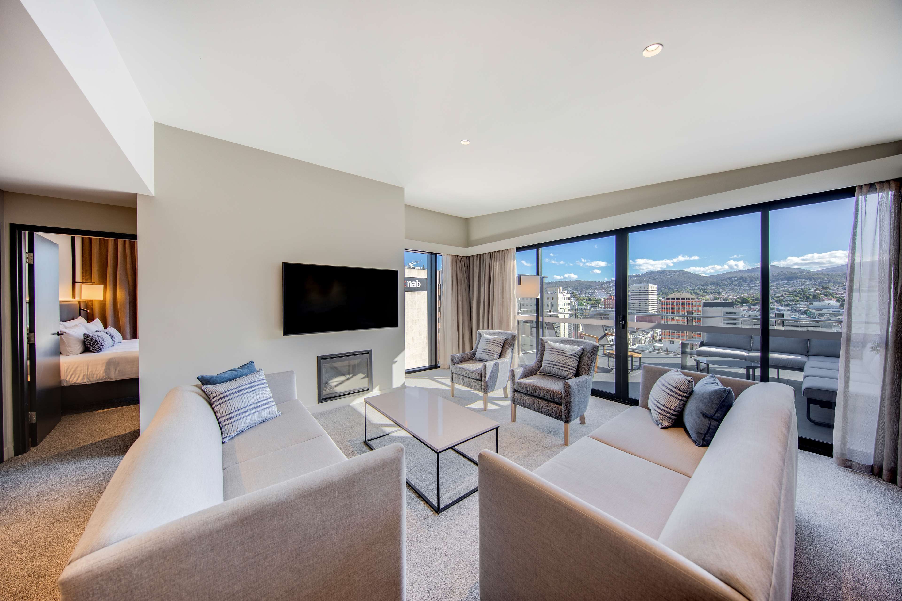 A modern hotel room with a large window offering a city view, a flat-screen TV mounted on the wall, and a cozy seating area with sofas and armchairs.