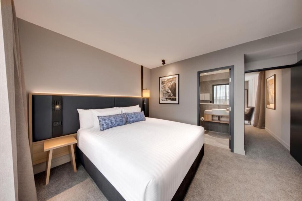 Une chambre d’hôtel moderne avec un grand lit, une table de chevet et une salle de bain visible à travers une porte ouverte.