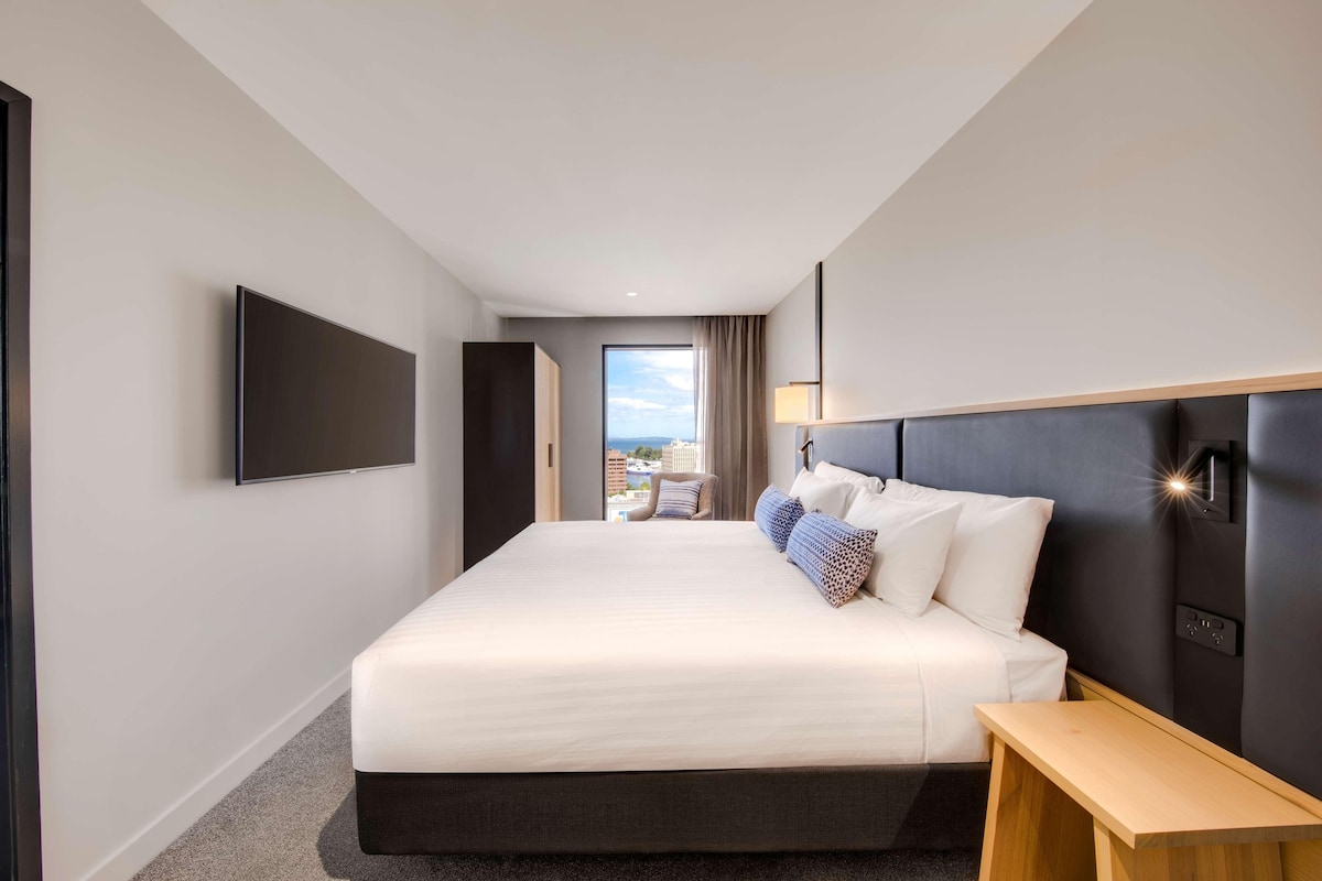 A modern hotel room with a large bed, a flat-screen TV mounted on the wall, a bedside table, and a view of the city through the window.