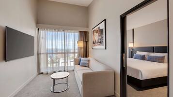 A hotel room with a sofa, a bed, a TV, a table, and a window with curtains.