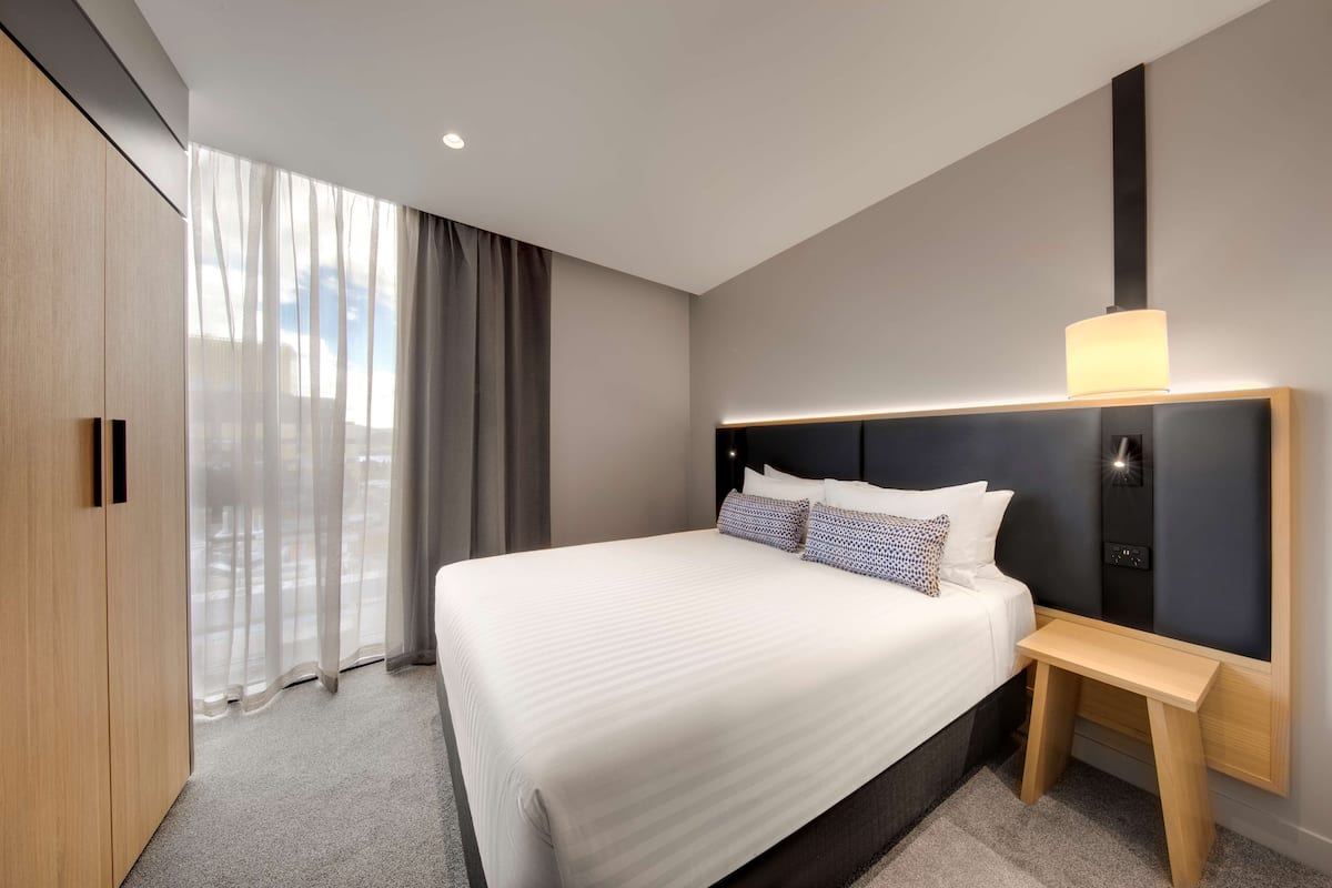 Moderni hotellihuone, jossa on suuri sänky, yöpöytä, sängynpääty, jossa on sisäänrakennettu valaistus, ja näkymä ohuiden verhojen läpi.