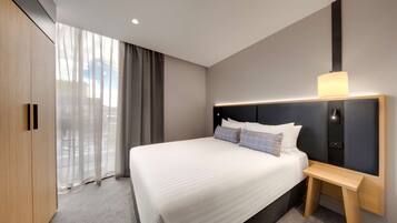 Quarto de hotel moderno com uma cama grande, mesa de cabeceira, cabeceira com iluminação integrada e vista através de cortinas translúcidas.