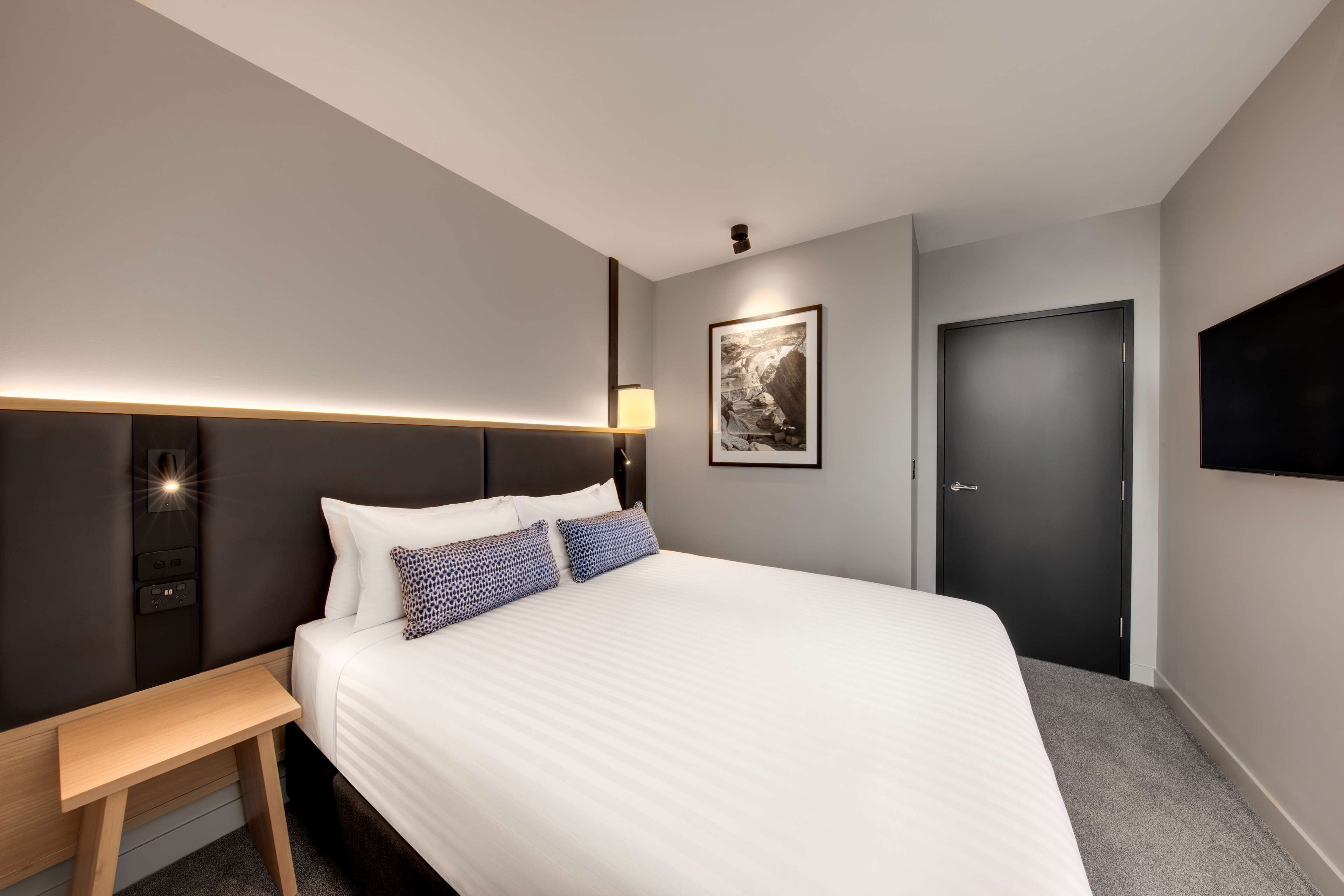 Moderni hotellihuone, jossa on suuri sänky, yöpöytä ja seinälle asennettu televisio.