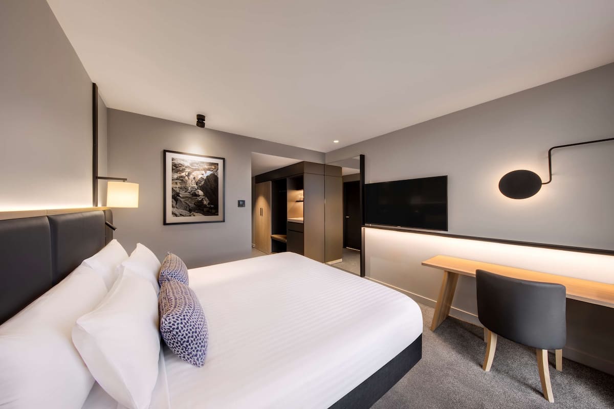 Moderni hotellihuone, jossa on suuri sänky, työpöytä ja tuoli. Seinälle on asennettu taulutelevisio, ja sängyn yläpuolella on kehystetty taulu.