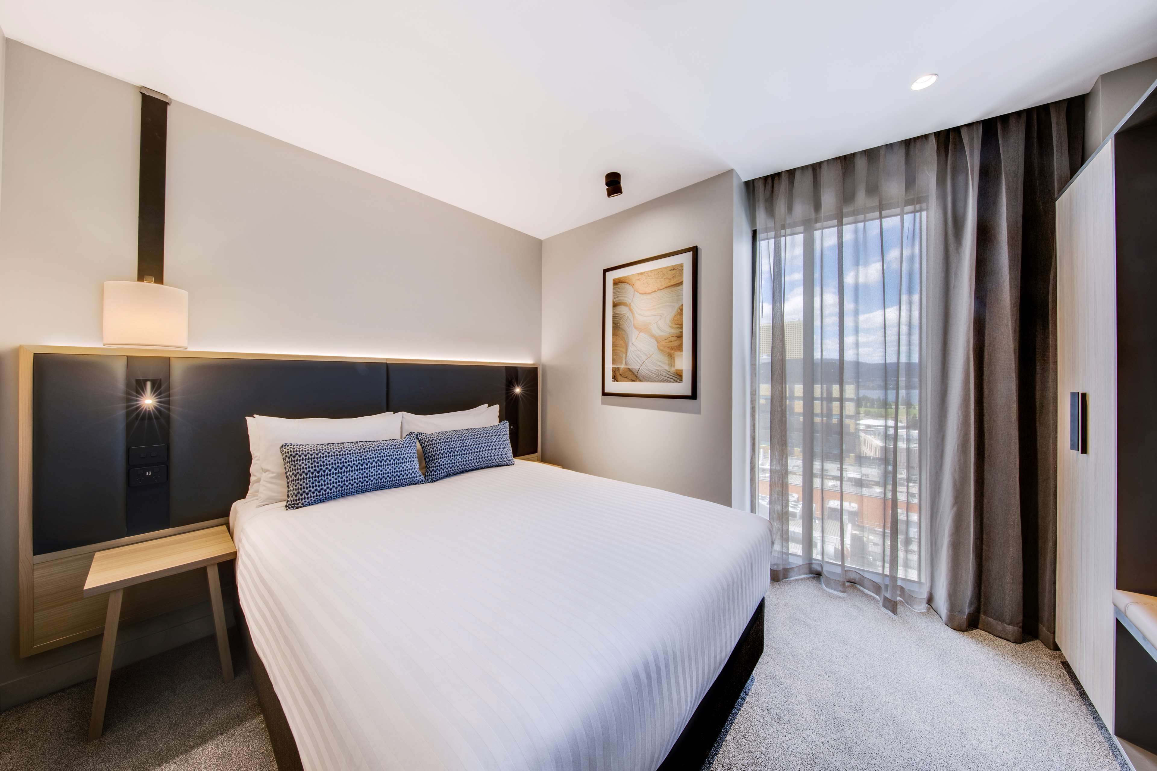 Une chambre d’hôtel moderne dotée d’un grand lit, d’une table de chevet, d’une commode, d’une tête de lit, d’une fenêtre avec des rideaux et offrant une vue sur la ville.