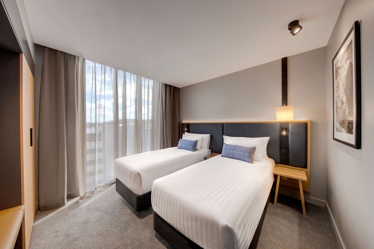 Ein Hotelzimmer mit zwei Betten, einem Nachttisch mit Lampe, einem gerahmten Bild an der Wand und einem Fenster mit Vorhängen.