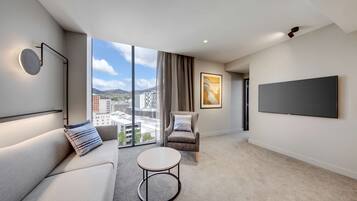 Una habitación de hotel moderna con un ventanal grande, un sofá, una silla, una mesita redonda pequeña y un televisor de pantalla plana montado en la pared.