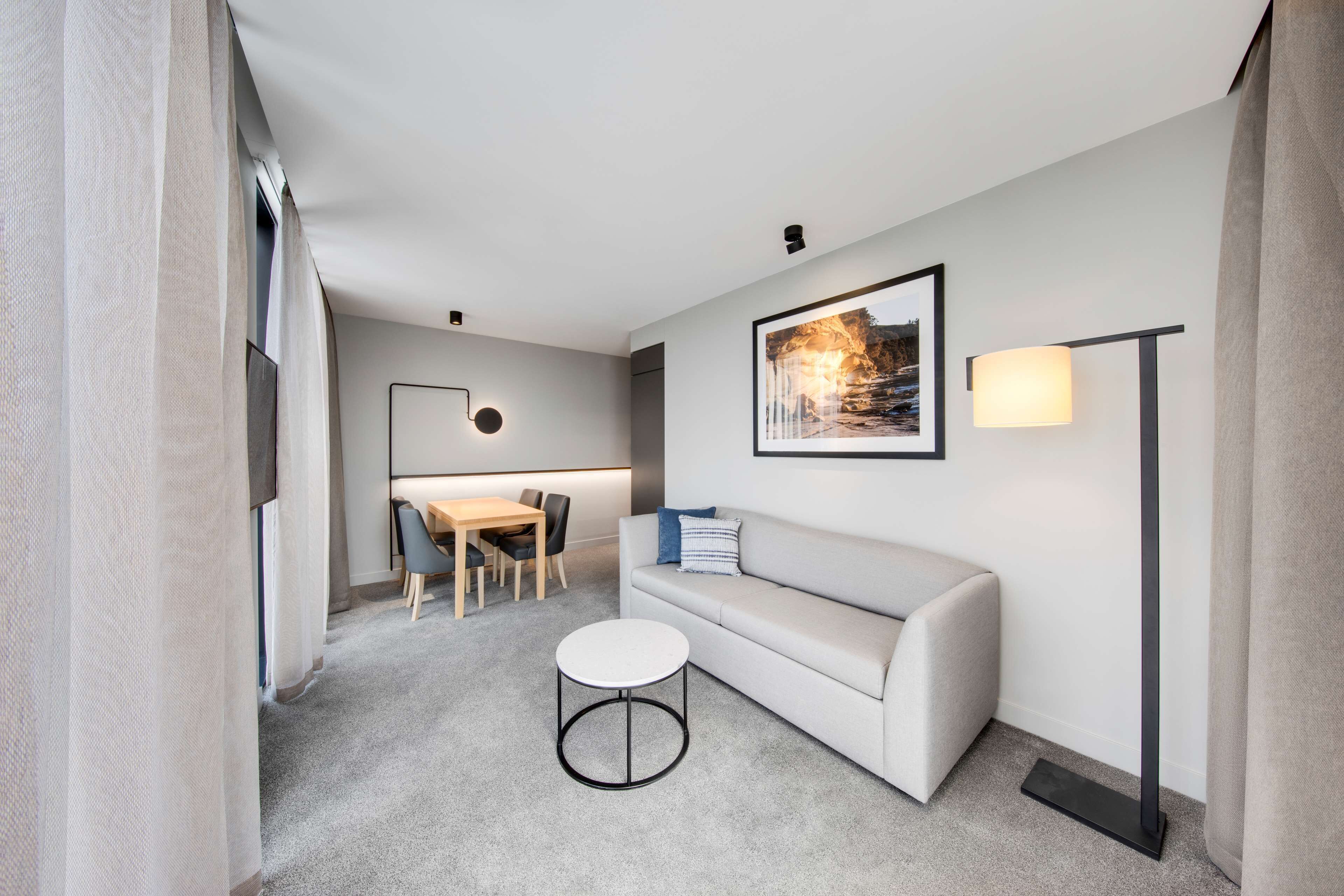 Una habitación de hotel moderna con un sofá gris, una mesita de centro redonda, un escritorio con sillas, un cuadro enmarcado en la pared y una lámpara de pie.