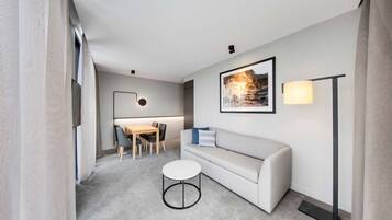 Una habitación de hotel moderna con un sofá gris, una mesita de centro redonda, un escritorio con sillas, un cuadro enmarcado en la pared y una lámpara de pie.