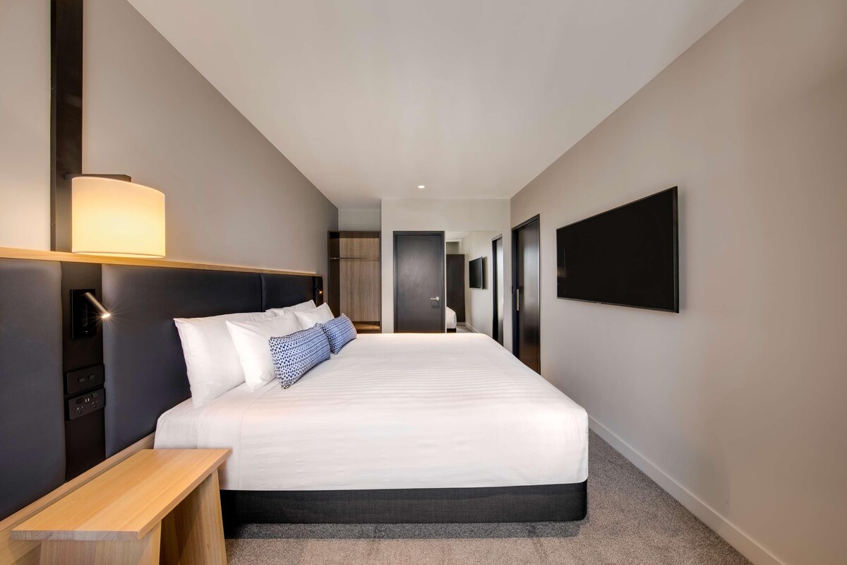 A modern hotel room with a large bed, bedside tables, a flat-screen TV, and a hallway leading to other rooms.